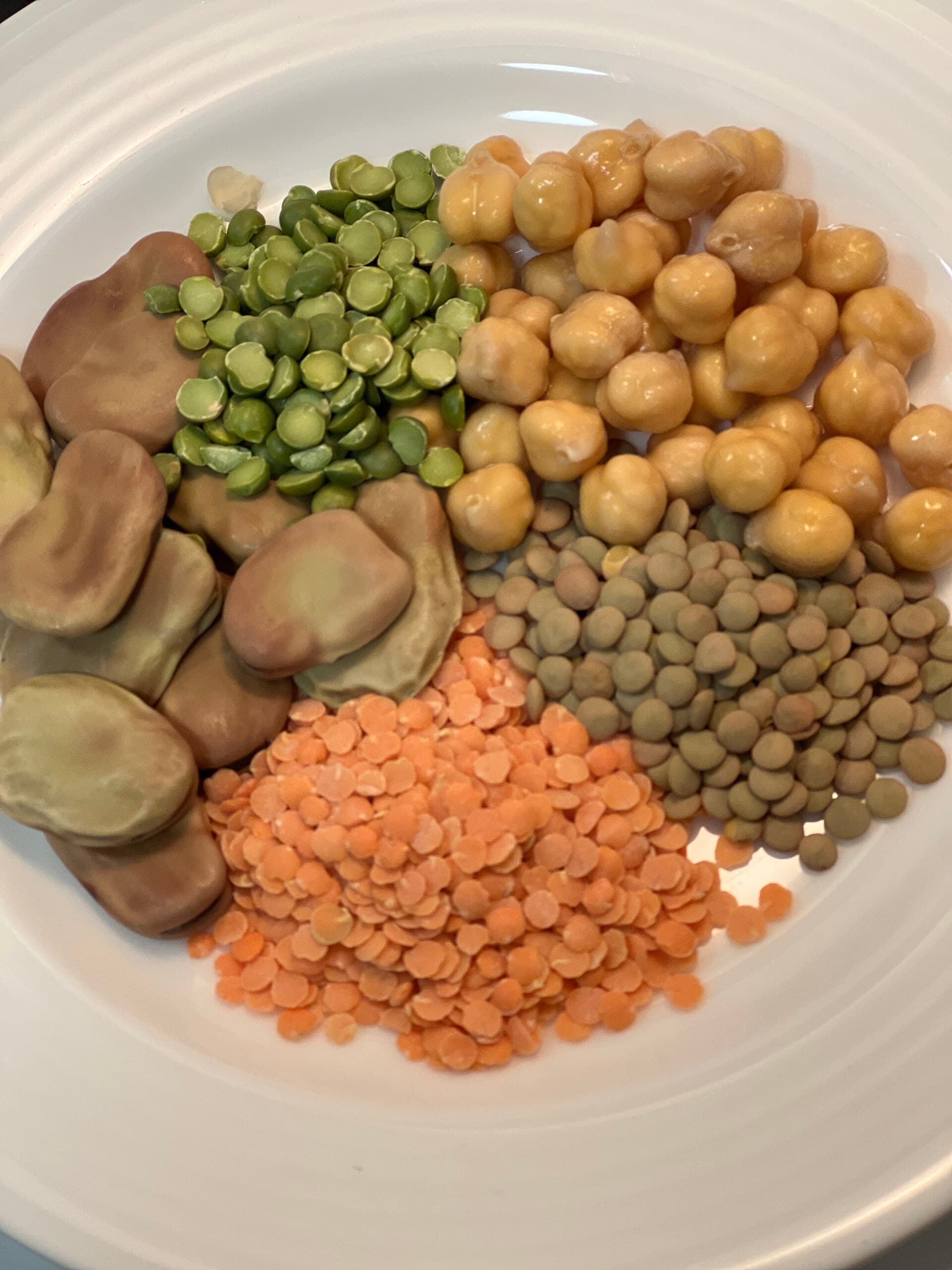 Assorted legumes on a white plate: fava beans, green split peas, chickpeas, brown and red lentils